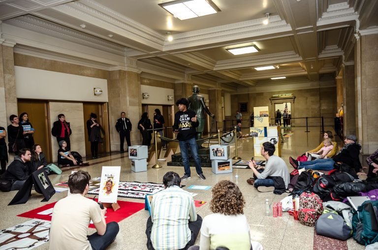 A speaker stands at the center of a crowd of seated demonstrators inside City Hall.