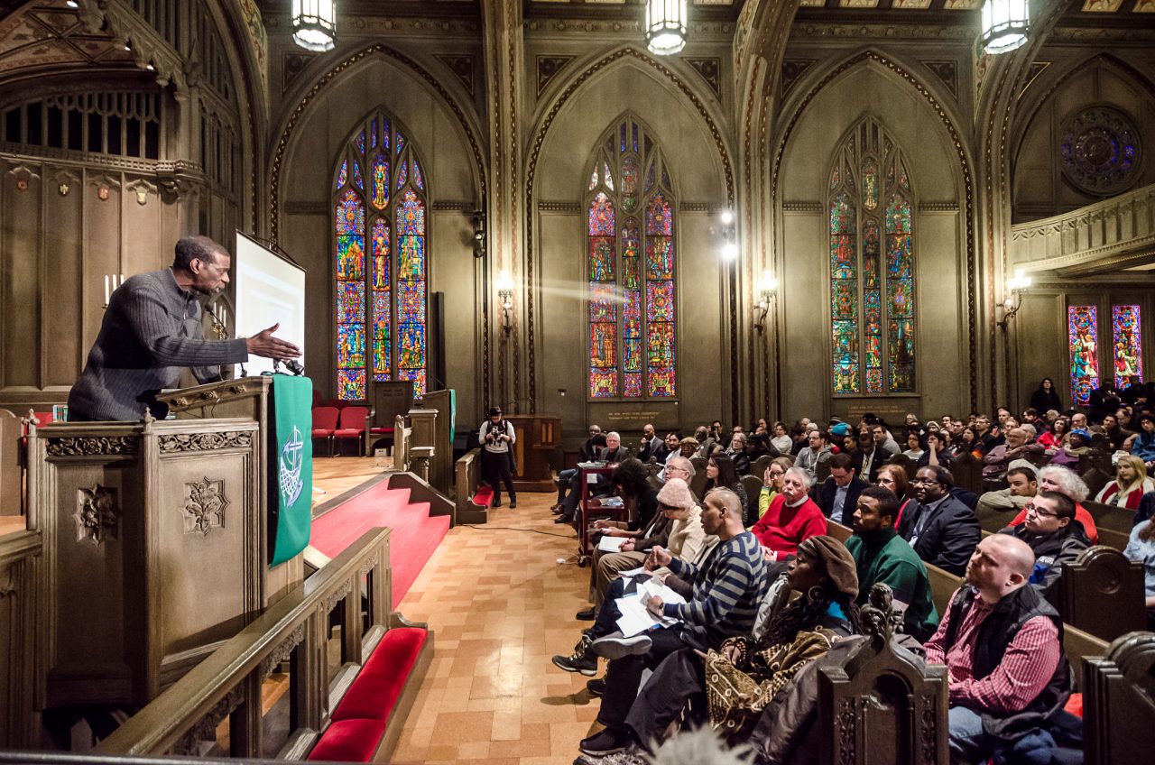 A speaker at the pulpit addresses a congregation seated in the temple, with large stained glass windows in the background.