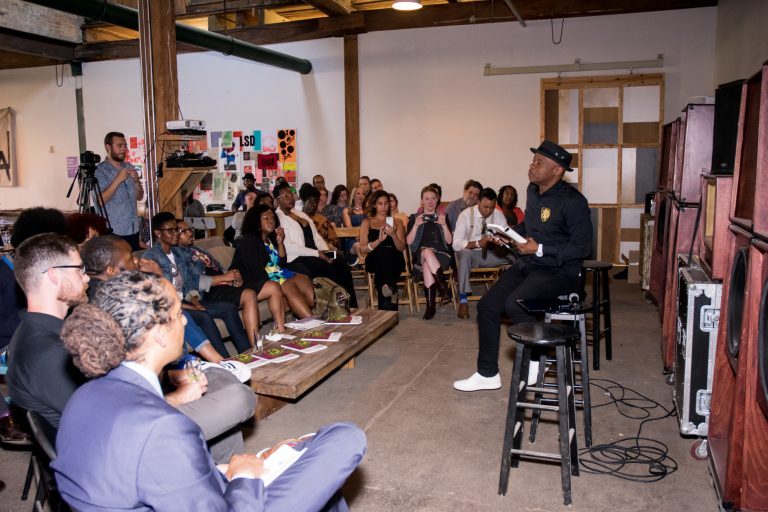 An audience of people listening to a presenter sitting on a stool and holding a book