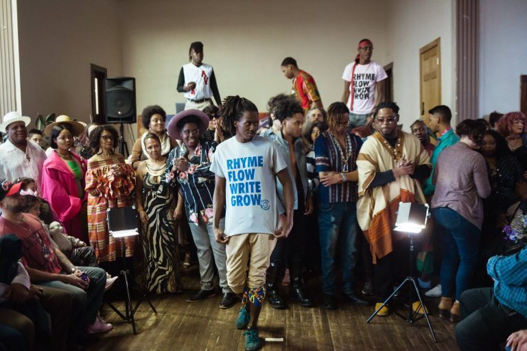 A crowd of people clapping. A man in front wears a shirt that reads "Rhyme / Flow / Write / Grow."