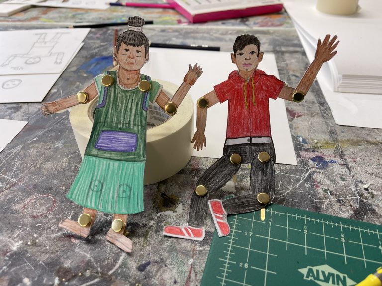 Two paper puppets, a boy and his grandmother, propped up on an artist's work table