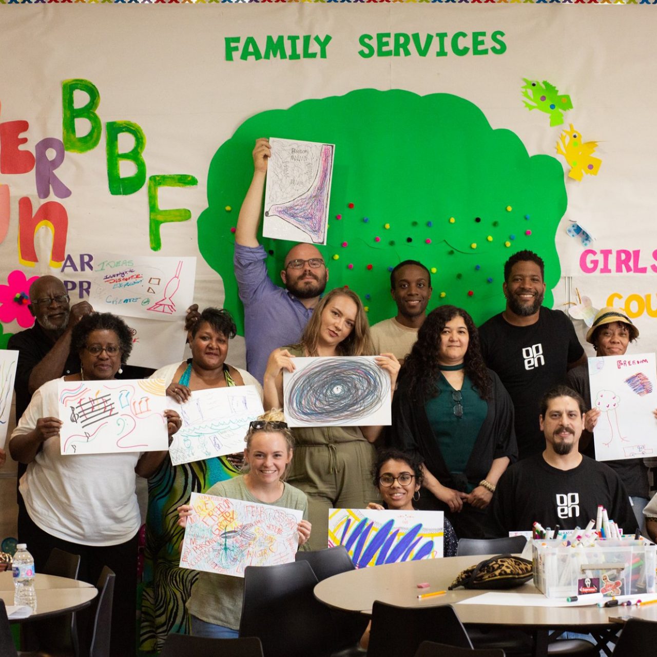A group of people holds up drawings and paintings in a classroom